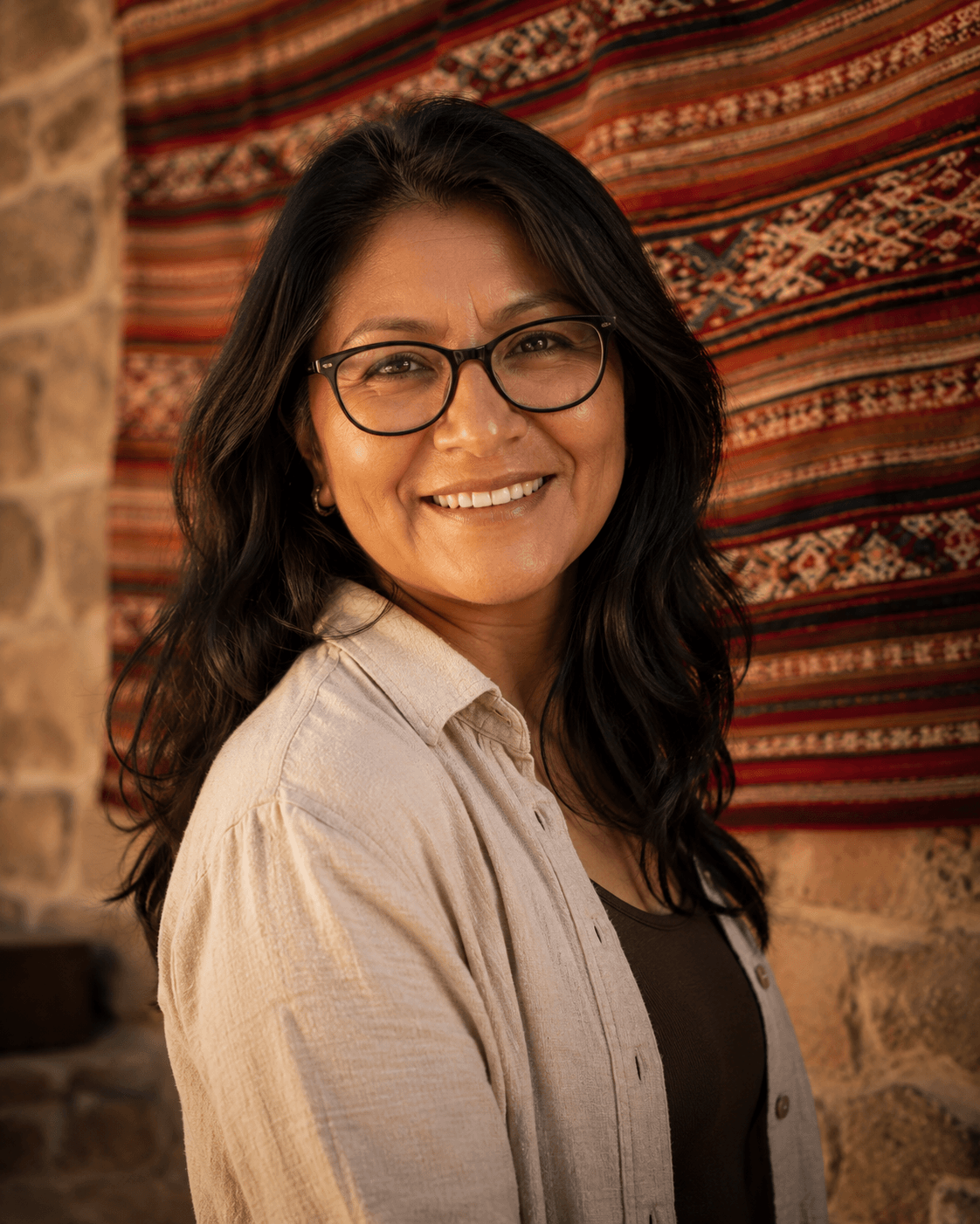 Portrait of La Illimani — Bolivian woman with glasses and a warm direct smile, in front of a woven aguayo wall hanging.